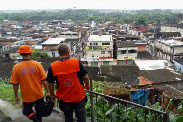 Imagens coloridas mostram dois homens de jaleco laranja da Defesa Civil observando cidade atingida pelas chuvas do alto de um morro