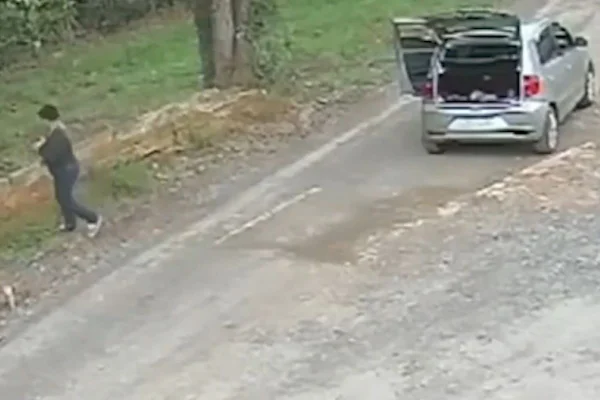 Imagem colorida mostra carro estacionado em estrada com poirta-malas aberto, mulher está caminhando em direção ao meio fio - Metrópoles