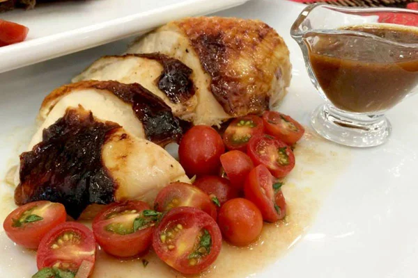 Na foto, um frango assado fatiado com tomates cereja e molho marrom em cima de um prato branco