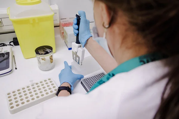 Mulher uniformizada em laboratório realizando testes com seringas em bancada, vista de costas - Metrópoles