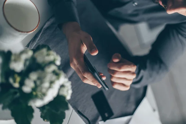 Imagem de um homem mexendo no celular sobre uma mesa com um buquê de flores e uma caneca