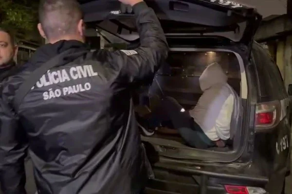 Imagem colorida de agente policial, com uniforme preto, abrindo porta-malas de carro; dentro, há um homem com blusa de moletom cinza, com capuz - Metrópoles