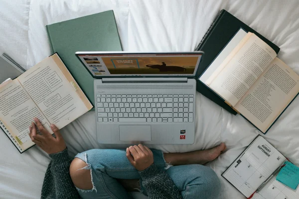 Imagem mostra mulher sentada em uma cama, cercada por livros abertos e um computador - Metrópoles