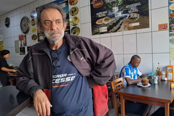 imagem colorida mostra foto de homem em restaurante. ele está com o braço apoiado em uma bancada, tem cabelo e barba grisalhos