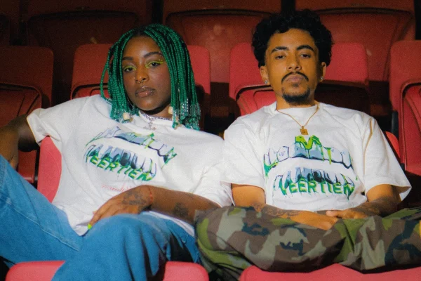 A imagem mostra duas pessoas sentadas em uma arquibancada usando a mesma camiseta branca
