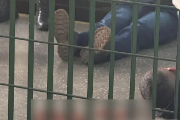 Imagem colorida de homem caído no chão baleado e outro sentado preso. Metrópoles