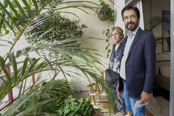 Imagem colorida mostra Ricardo Nunes, homem branco, de cabelo e barba pretos, vestindo terno azul, camisa branca e calça jeans, em uma sacada de prédio decorada com plantas - Metrópoes