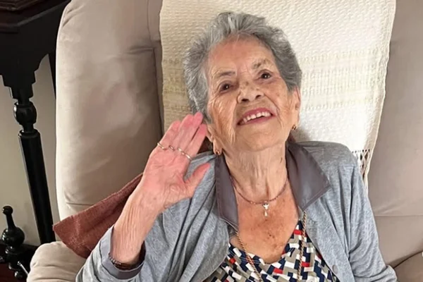 Foto colorida de mulher sentada em um sofá e acenando - Metrópoles