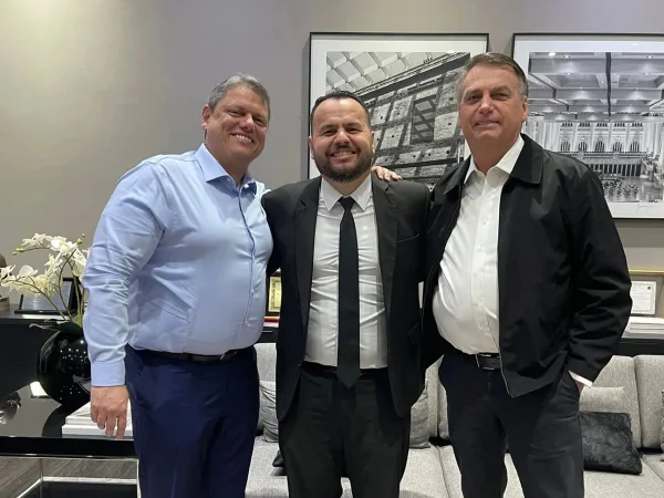 Imagem colorida mostra Tarcísio de Freitas, homem branco de camisa azul, Gil Diniz, de terno e gravata, e Jair Bolsoanro, de jaqueta preta e camisa branca. Os três estão abraçados, virados para a câmera, em uma sala de tons pasteis - Metrópoles