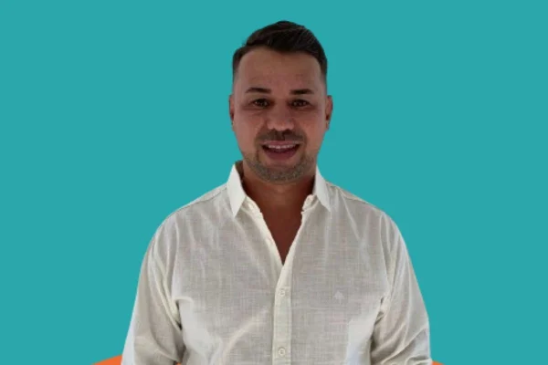 Homem branco de cabelos curtos e camisa branca com o primeiro botão aberto; fundo azul/ Metrópoles