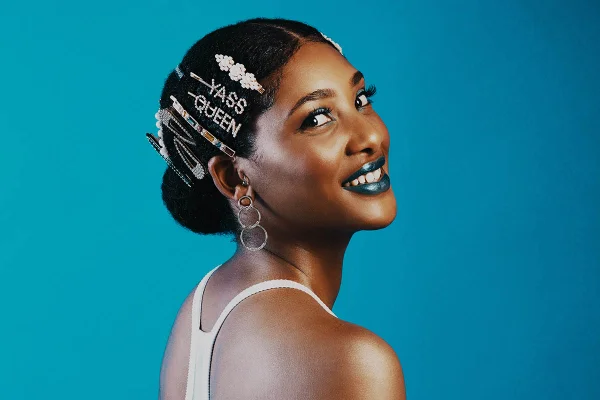 Mulher negra com cabelo preso e várias presilhas enfeitando a cabeça. Ela está em um fundo azul e usa o batom da mesma cor.