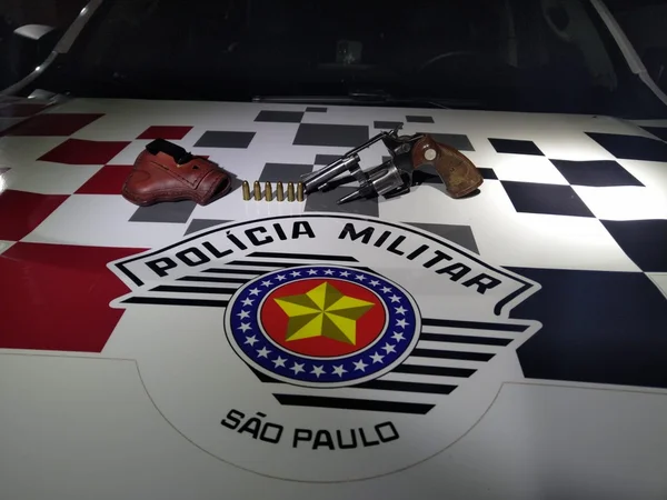 Imagem colorida mostra arma apreendida pela PM em cima de um capô de carro com o logo da Polícia Militar; homem que se diz proprietário do objeto foi preso em flagrante - Metrópoles