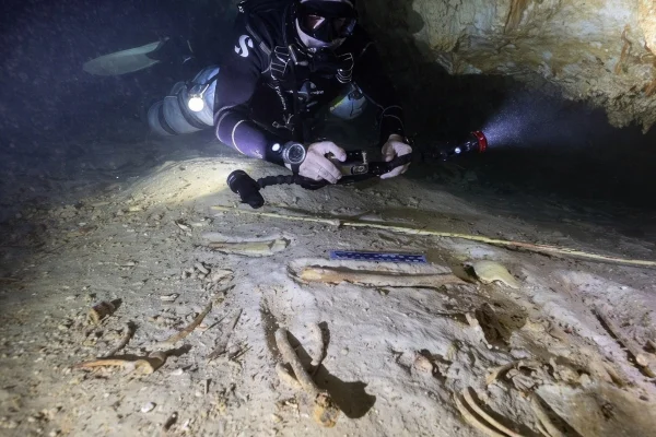 Foto subaquática de nadador com câmera iluminnando e tirando foto de esqueletos humanos no fundo de uma caverna - Metrópoles.