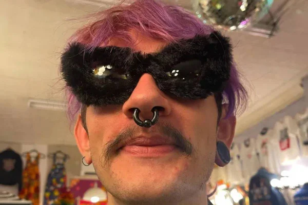 Homem de óculos escuros, bigode, piercing e cabelo rosa - Metrópoes