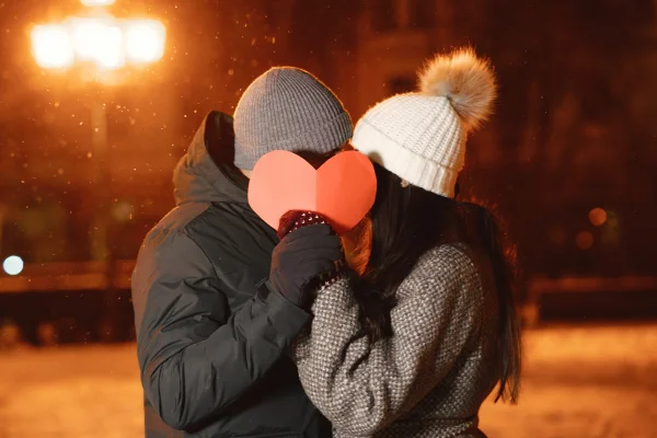 Imagem colorida com casal segurando coração na frente do rosto