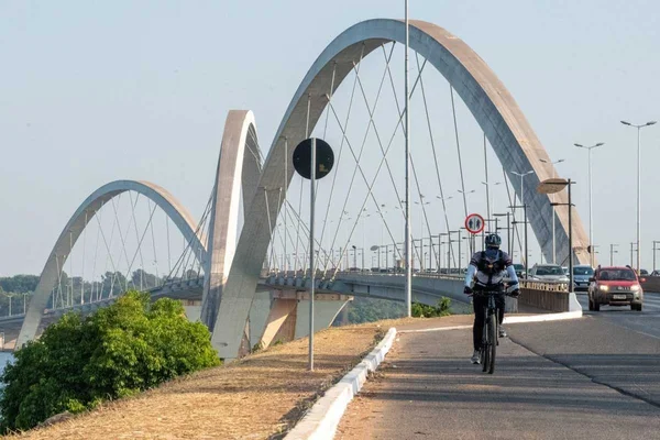 Ciclista pedala, com capacete e casaco azul e preto, com Ponte JK ao fundo. Há um arbusto à esquerda, veículos passando pela via, uma placa de trânsito ao fundo, além de postes ao longo da ponte