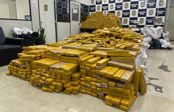 Embalagens em cor alaranjadas empilhadas em chão de sala com logotipo da Polícia Civil ao fundo
