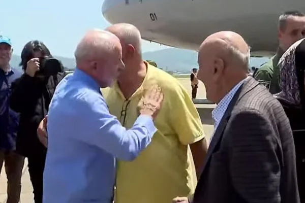 Três homens interagem ao lado de avião, em pista de pouso, em dia ensolarado - Metrópoles