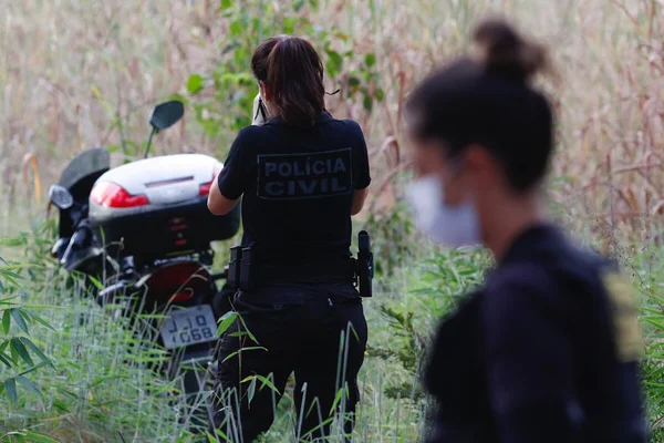 Mulher de máscara com cabelo preso e blusa preta com o rosto de perfil desfocado. Ao fundo, uma mulher de costas com roupa preta e rabo de cavalo ao lado de motocicleta em meio a matagal