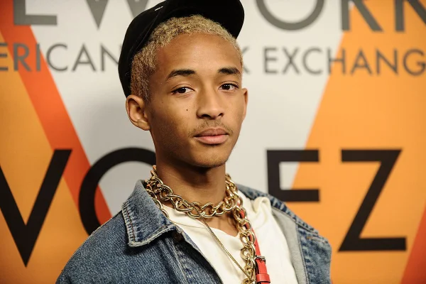 Ator e cantor Jaden Smith, um rapaz negro com cabelo raspado louro, posando para foto em um fundo branco no evento da Louis Vuitton. Ele usa uma camiseta branca, jaqueta jeans e