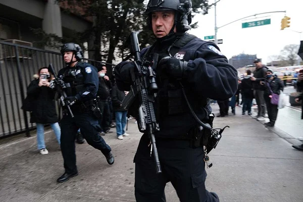 Policiais fortemente armados, em uniforme preto, passam pela rua próximo a atentado em metrô, onde suspeito atirou em pessoas, Nova Iorque. Ao redor deles, forma-se uma aglomeração de curiosos - Metrópoles