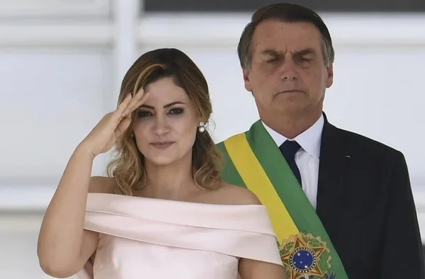 Na imagem colorida, um homem e uma mulher estão posicionados no centro da imagem. A mulher usa roupa rosa e o homem, atrás dela, usa terno preto, camiseta branca e está com a faixa presidencial
