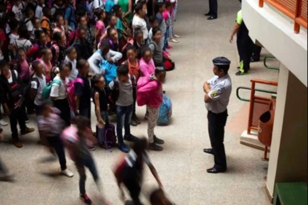 imagem colorida mostra adolescentes em escola usando uniforme em frente a militar da PM, que usa farda