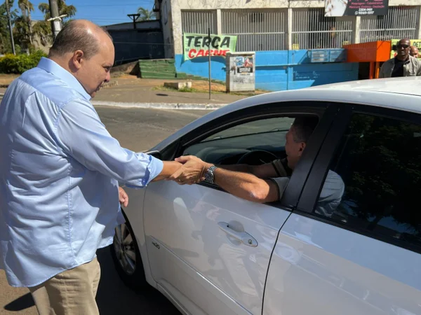 Homem branco de blusa social azul, calvo, apertando a mão de um homem branco dentro de um carro branco