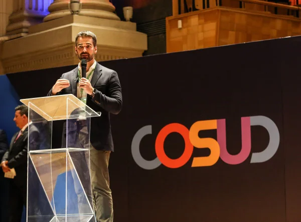 Imagem colorida mostra Eduardo Leite, homem branco, de cabelo e barba aparada castanhos, paletó azul, camisa branca e calça verde musgo, falando ao microfone em um púlpito transparente, em um palco, com um painel escrito "Cosud"com letras coloridas ao fundo - Metrópoles