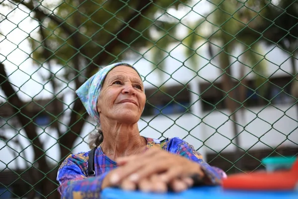 Mulher idosa com touca azul na cabeça, blusa azul de manga comprida e com uma mão em cima da outra. Ela está com o rosto para cima, em frente a uma grade