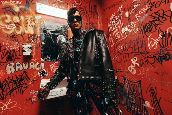 Fotos de campanha da coleção da Tommy Hilfiger com o estilista Richard Quinn. Um homem jovem e de pele branca está em um banheiro vermelho e usa uma roupa xadrez preta e vermelha e uma jaqueta de couro.