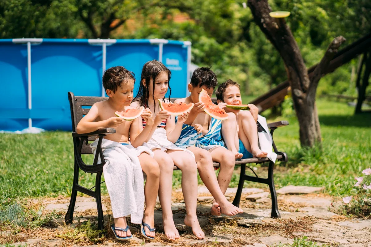 Imagem colorida de crianças comendo melancia perto de uma piscina - Metrópoles