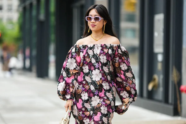 Mulher jovem e de pele branca, de origem asiática, com cabelos lisos e longos, andando pelas ruas de Nova York durante a semana de moda na cidade. Ela usa um vestido florido com fundo preto, óculos escuros de armação lilás e uma bolsa de couro branca.