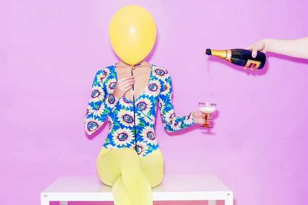 Mulher branca e jovem posa para foto em um fundo rosa. Ela usa um maiô azul com flores de girassol estampadas e uma meia calça amarela. Com uma das mãos segura um balão amarelo e, com a outra, uma taça com champanhe. - Metrópoles