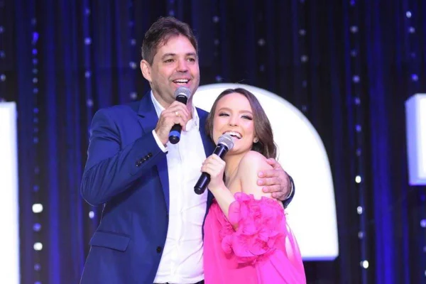 Marcelo Germano, homem de meia idade com camisa branca e blazer azul escuro, abraçado em Larissa Manoela, que veste vestido rosa. Os dois seguram e falam em microfones - metrópoles