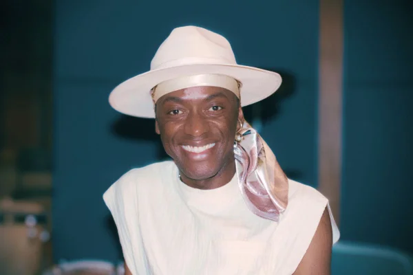 imagem colorida mostra cantor jota.pê sorrindo. ele é negro, usa um chapéu branco e uma regata da mesma cor. abaixo do chapéu tem um lenço estampado de cor rosa claro.
