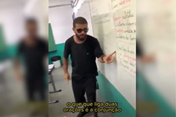imagem colorida mostra professor apontando para lousa. ele veste preto e usa óculos escuros