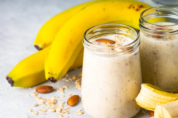 Foto colorida de copo com vitamina de banana. Ao fundo, há três frutas - Metrópoles
