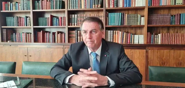 O presidente Bolsonaro durante fala em gabinete presidencial para vídeo. Ele está sentado e atrás é possível ver uma biblioteca cheia de livros nas estantes - Metrópoles