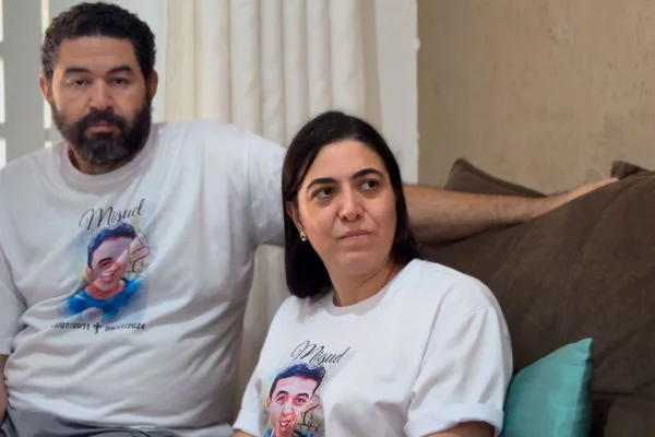 Pais de Miguel Fernandes Brandão vestem um camisa branca com a foto o filho. Fábio Brandão é um homem de 43 anos, branco, com barba. Genilva Brandão é uma mulher de 40 anos, branca, cabeço liso.