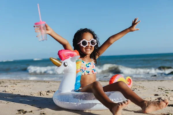 Foto colorida de menina na praia sentada em boia de unicórnio - Metrópoles