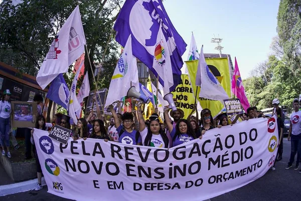 Imagem colorida mostra grupo de estudantes segurando uma faixa branca com letras pretas com a frase "Pela revogação do novo ensino médio e em defesa do orçamento". As pessoas ao fundo seguram várias bandeiras de entidades estudantis - Metrópoles