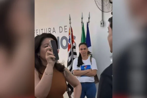 Imagens coloridas mostram mulher branca, cabelos pretos e longos, mostrando celular para homem durante discussão em prefeitura. De frente, uma mulher observa a cena