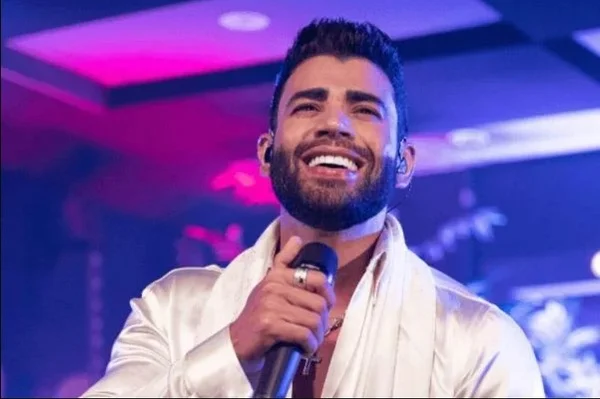 Gusttavo Lima cantor sertanejo em show. Ele usa roupa branca e tem um microfone na frente da boca