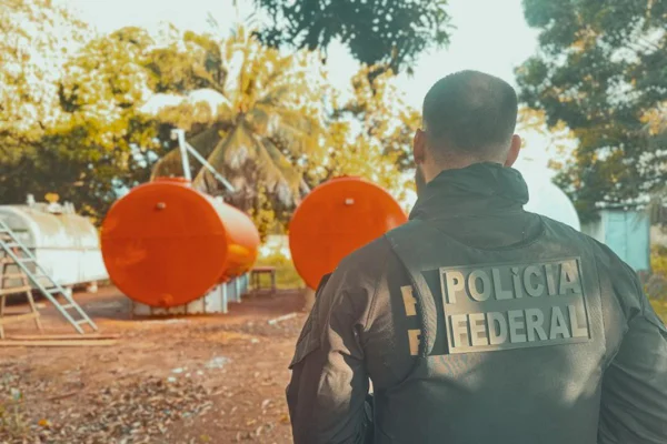 Foto colorido de agente da PF. Ao fundo galões de gasolina ilegal - Metrópoles