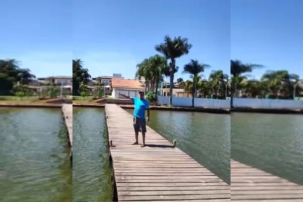 Imagem colorida de um homem de camiseta e bermuda azuis, apontando com o braço direito para a esquerda. Ele está sobre uma ponte no Lago Paranoá