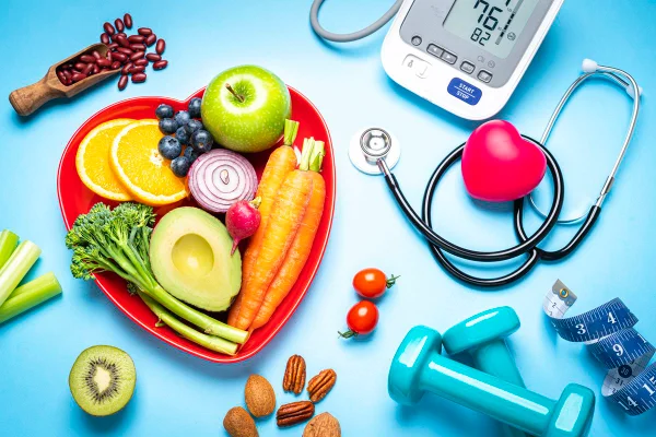 Foto colorida de superfície azul com estetoscópio, frutas dentro de um prato em formato de coração, equipamento para medir pressão arterial, peso para fazer exercícios e fita métrica - Metrópoles