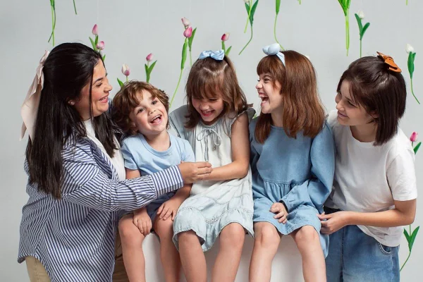 Foto colorida de mulher com um menino, duas meninas e uma adolescente. Ao fundo do cenário, tem flores suspensas - Metrópoles