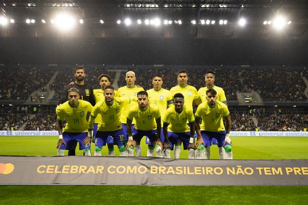 Jogadores da seleção do Brasil em pose de formação para foto em campo, com arquibancada de estádio ao fundo. Eles usam camisa amarela e short azul - Metrópoles