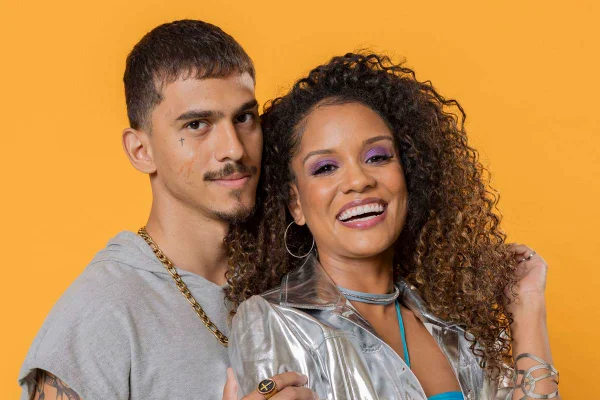 homem moreno de bigode posa com meio sorrido de camiseta cinza ao lado de mulher morena de cabelos cacheados que posa sorrindo com blusa furtacor - metrópoles